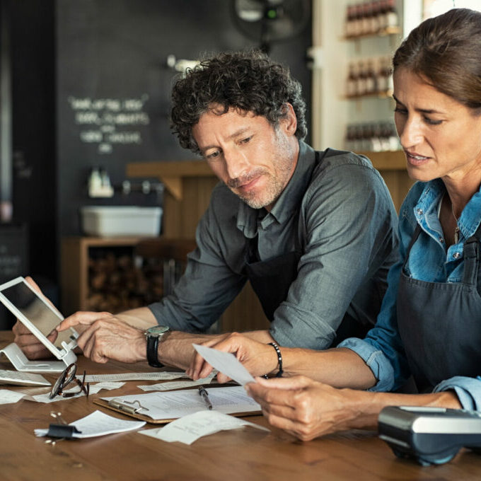 Man and woman sitting in cafeteria discussing finance for the month. Stressed couple looking at bills sitting in restaurant wearing uniform apron. Café staff sitting together looking at expenses and bills.